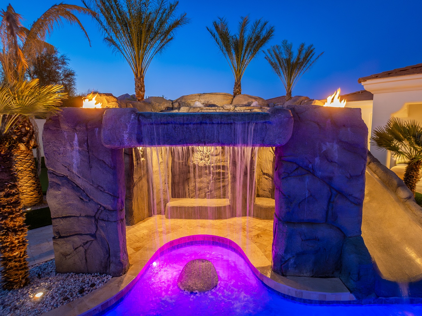 A stunning water feature glows under twilight skies, framed by illuminated palms and fire accents—your private desert oasis awaits with dramatic ambiance and luxury.