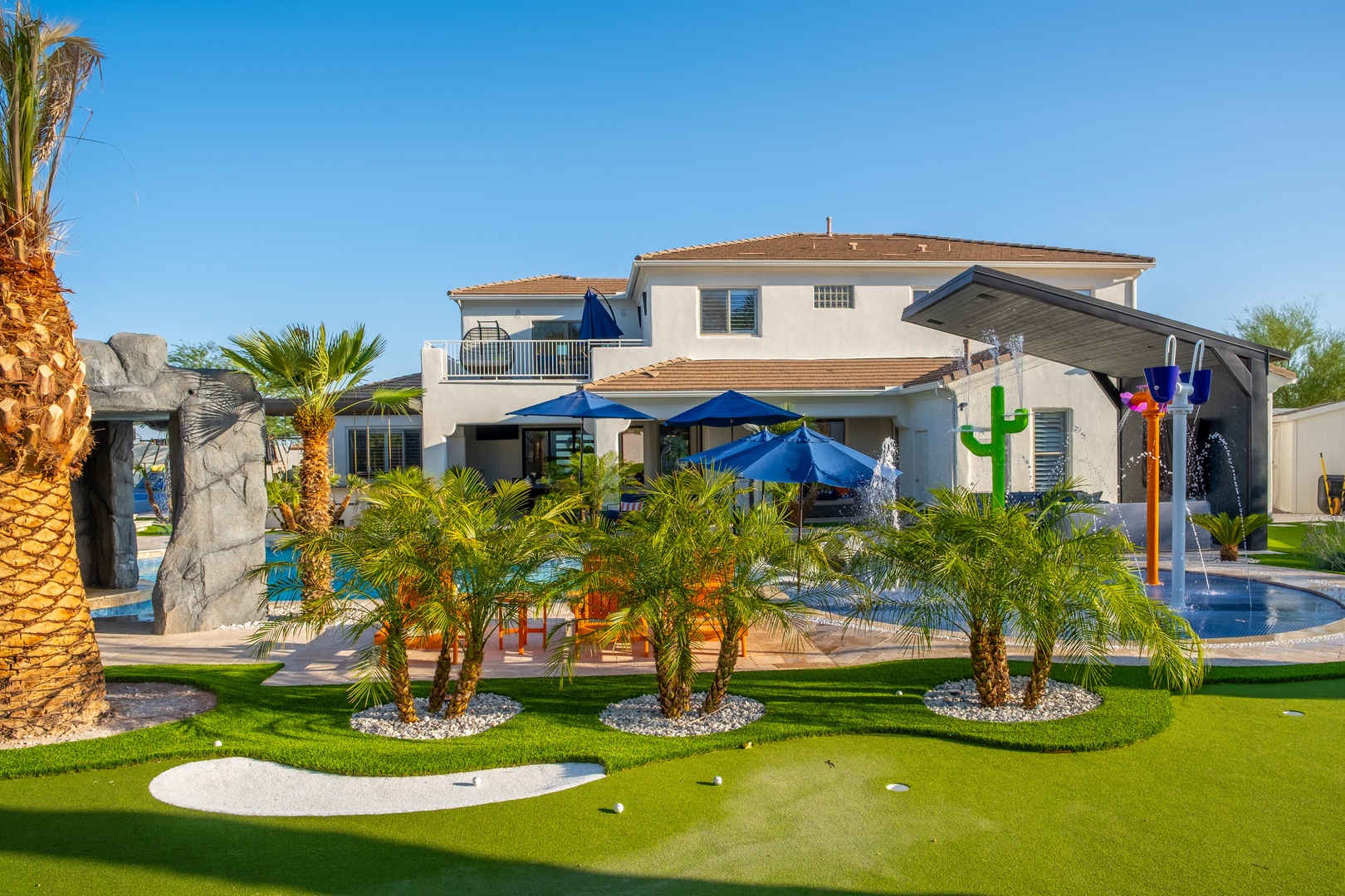 A striking vacation rental featuring a private resort-style backyard with palm trees, miniature golf green, water features, and multiple pool areas under sunny desert skies.