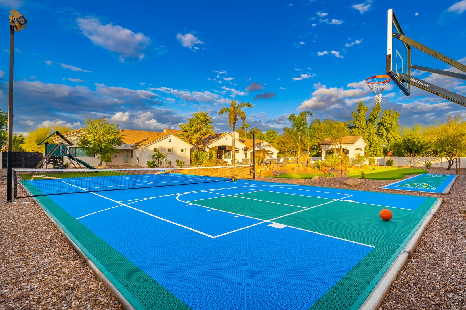 Outdoor recreation area featuring multi-sport court with basketball hoop, playground, and spacious grounds surrounded by palm trees under sunny skies.