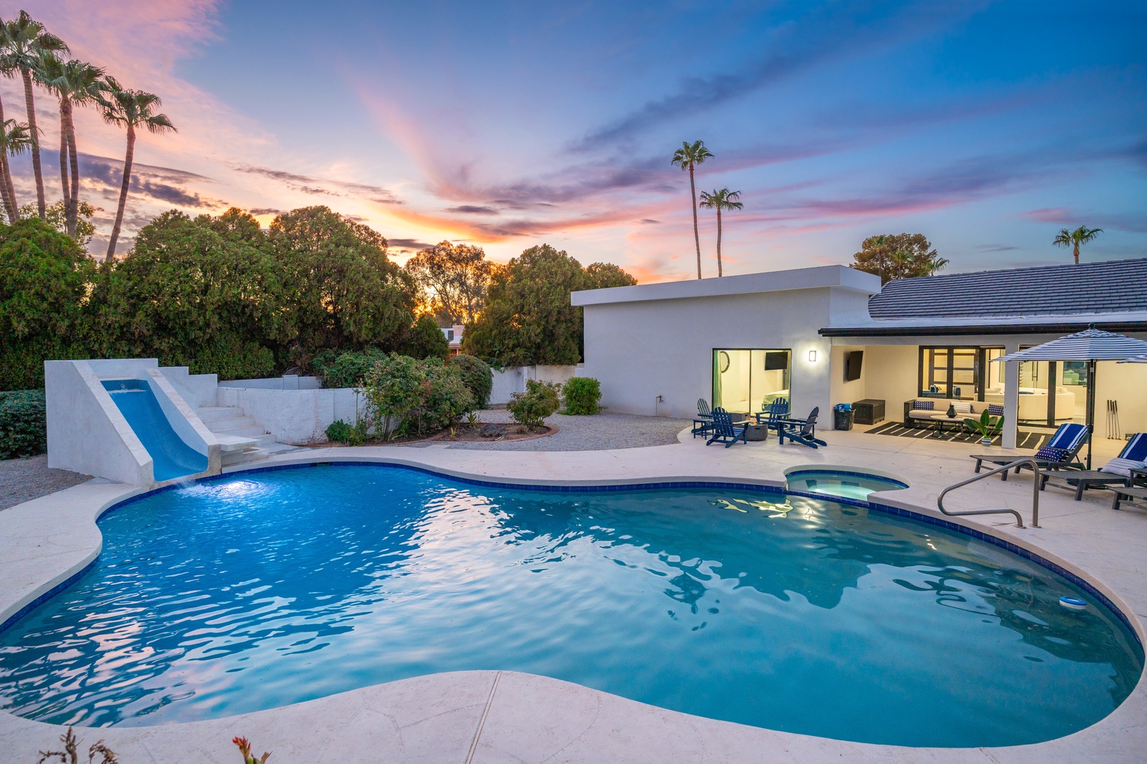 The property features a spacious pool with a slide, surrounded by mature palm trees and desert landscaping, with a covered patio area perfect for evening relaxation under painted sunset skies.