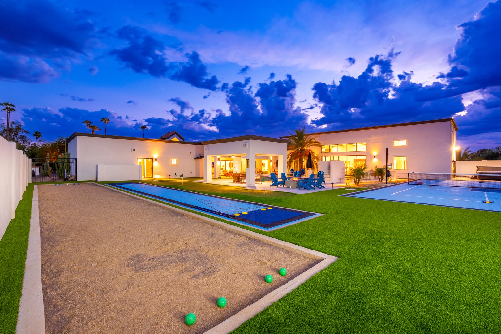 A modern desert retreat featuring bocce ball courts and sleek lap pools under evening skies, framed by manicured lawns and palm trees beyond the white stucco architecture.