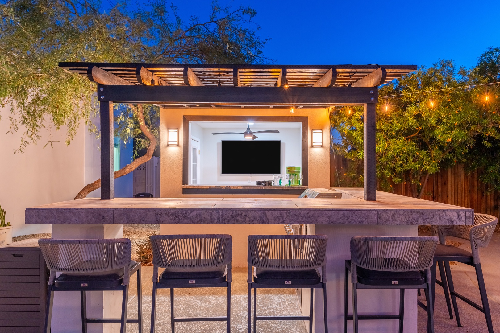 Picture yourself gathered at this sleek outdoor bar under twinkling lights, cocktails in hand, with a big screen for movie nights and lush greenery creating your private oasis.