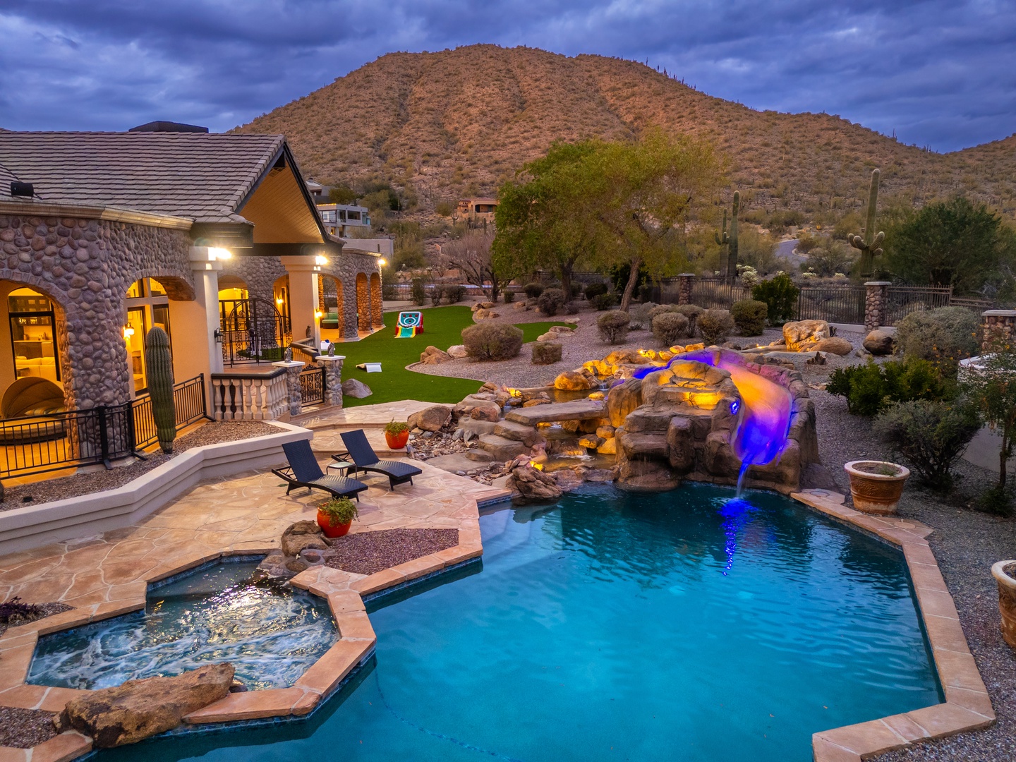 A stunning desert retreat features a sparkling pool with illuminated waterfall, adjacent hot tub, and dramatic mountain backdrop—your private oasis for unforgettable Arizona evenings under painted skies.