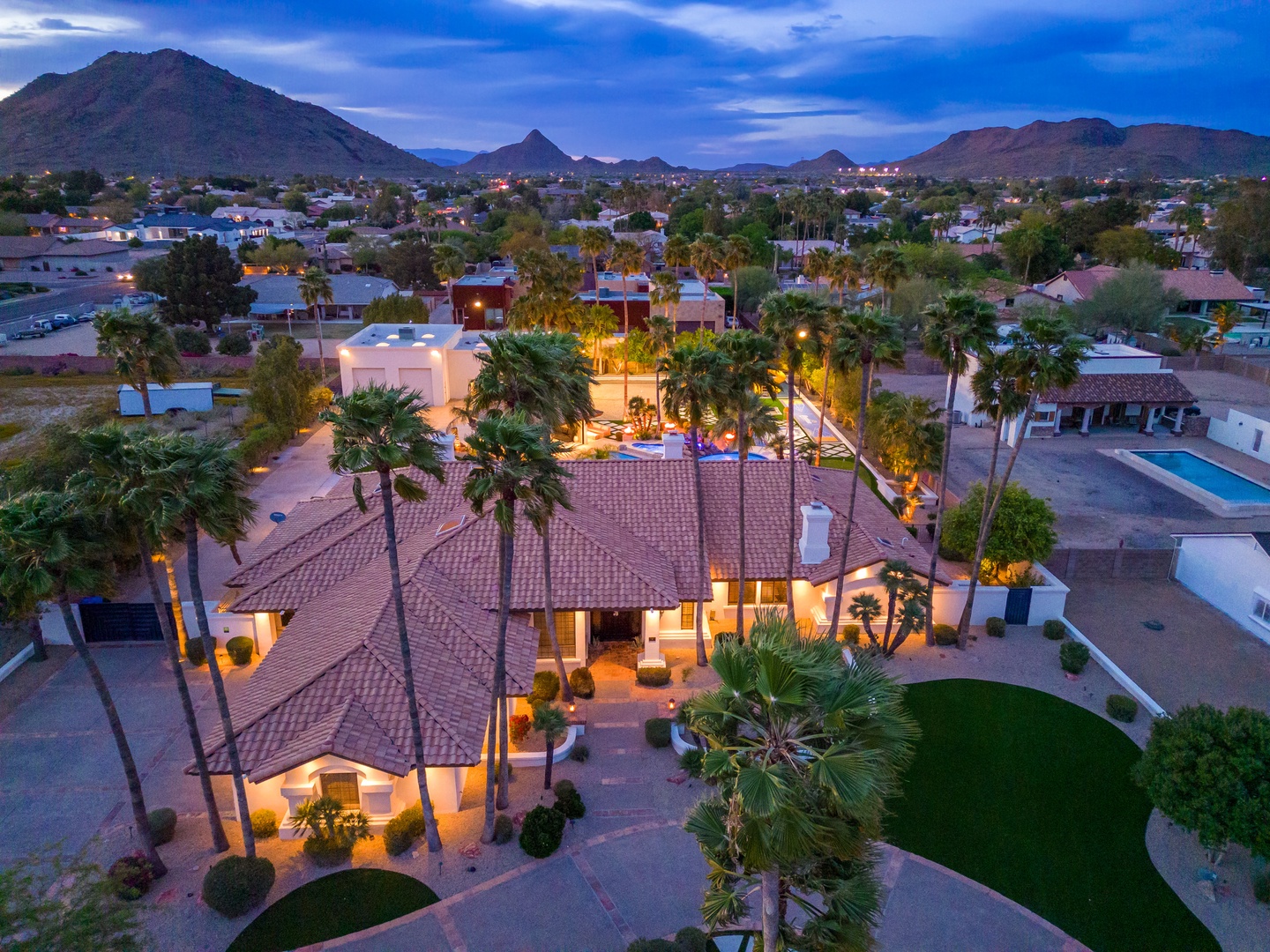 Aerial view of a desert estate featuring Spanish-style tile roofing, towering palms, and manicured lawns set against dramatic mountain peaks at twilight.
