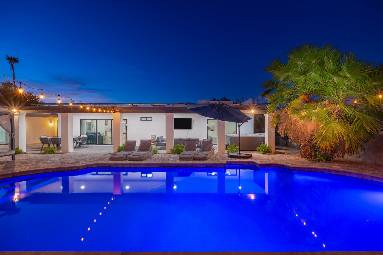 The property's inviting pool glows under twilight skies, surrounded by comfortable loungers, lush palms, and warm string lighting—a perfect desert oasis for evening relaxation.