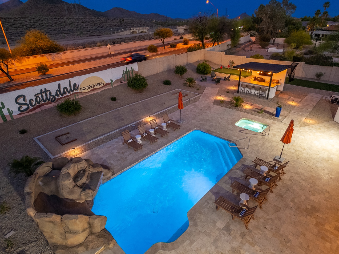 Aerial view of Scottsdale Daydreaming's beautifully lit pool area at twilight, featuring resort-style loungers, covered patio, and mountain backdrop.