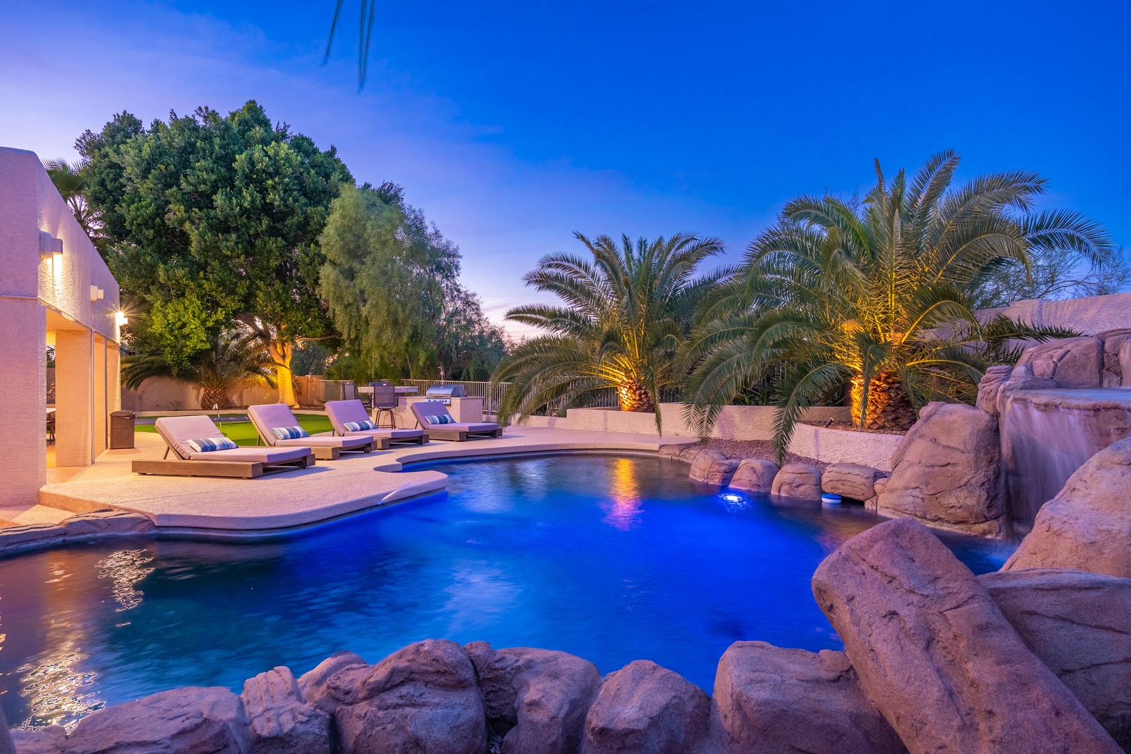 Luxurious resort-style pool features a dramatic waterfall flowing over natural rock formations. Palm trees frame the illuminated waters at twilight, while poolside loungers await under desert skies.