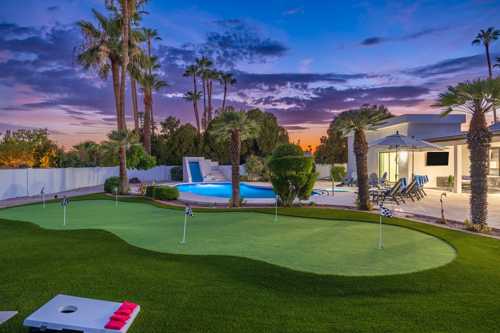 A stunning desert oasis featuring a private putting green, illuminated pool with slide, and covered patio beneath towering palms as twilight paints the sky in vibrant hues.