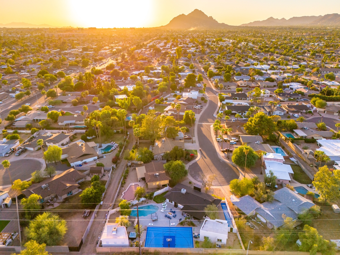 Aerial view captures the property nestled in a peaceful residential neighborhood with dramatic mountain peaks rising in the distance. The area features tree-lined streets and nearby homes bathed in warm golden light.