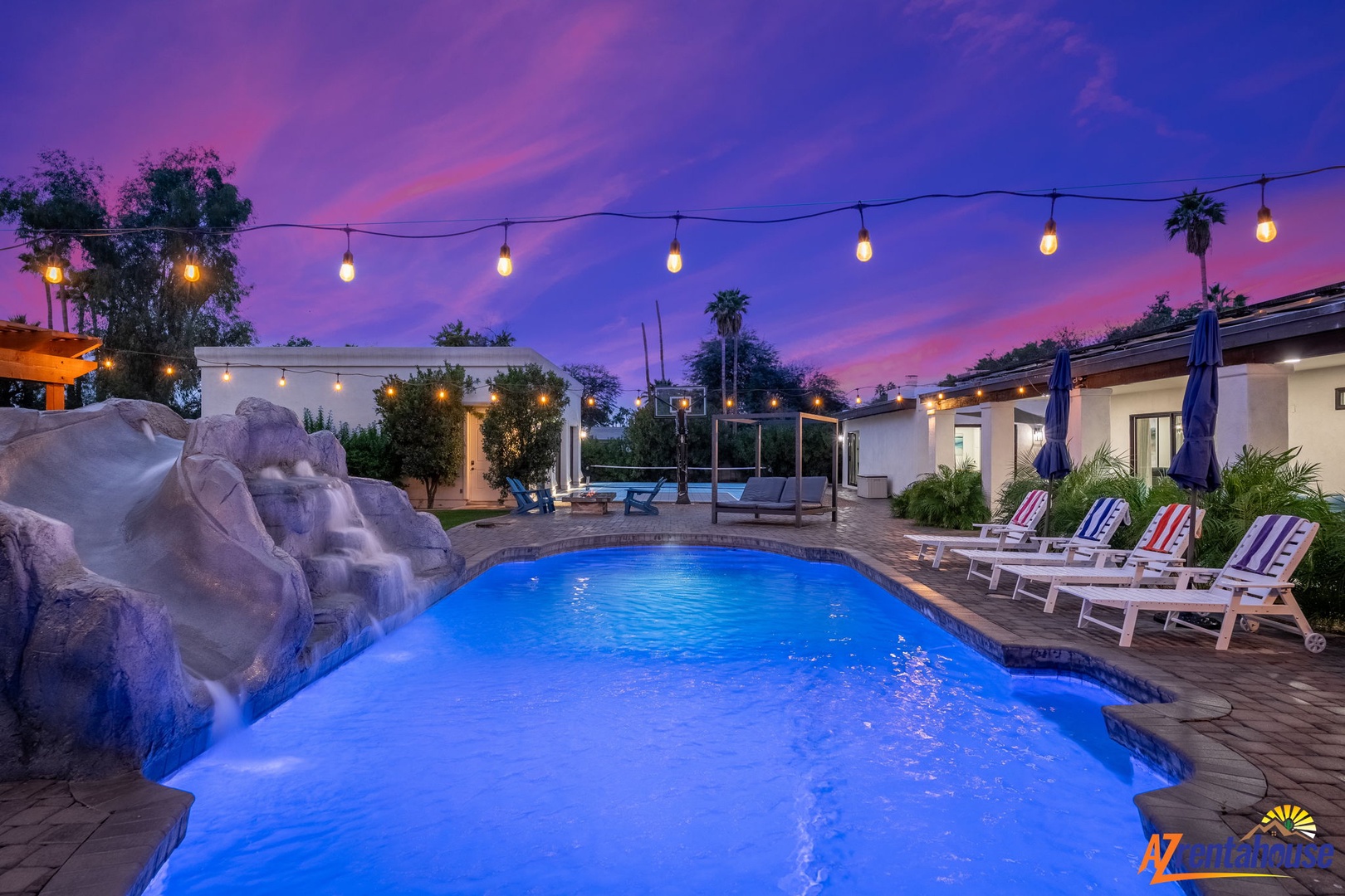 A stunning desert oasis featuring pool lighting and waterfall under vibrant purple-pink evening skies.