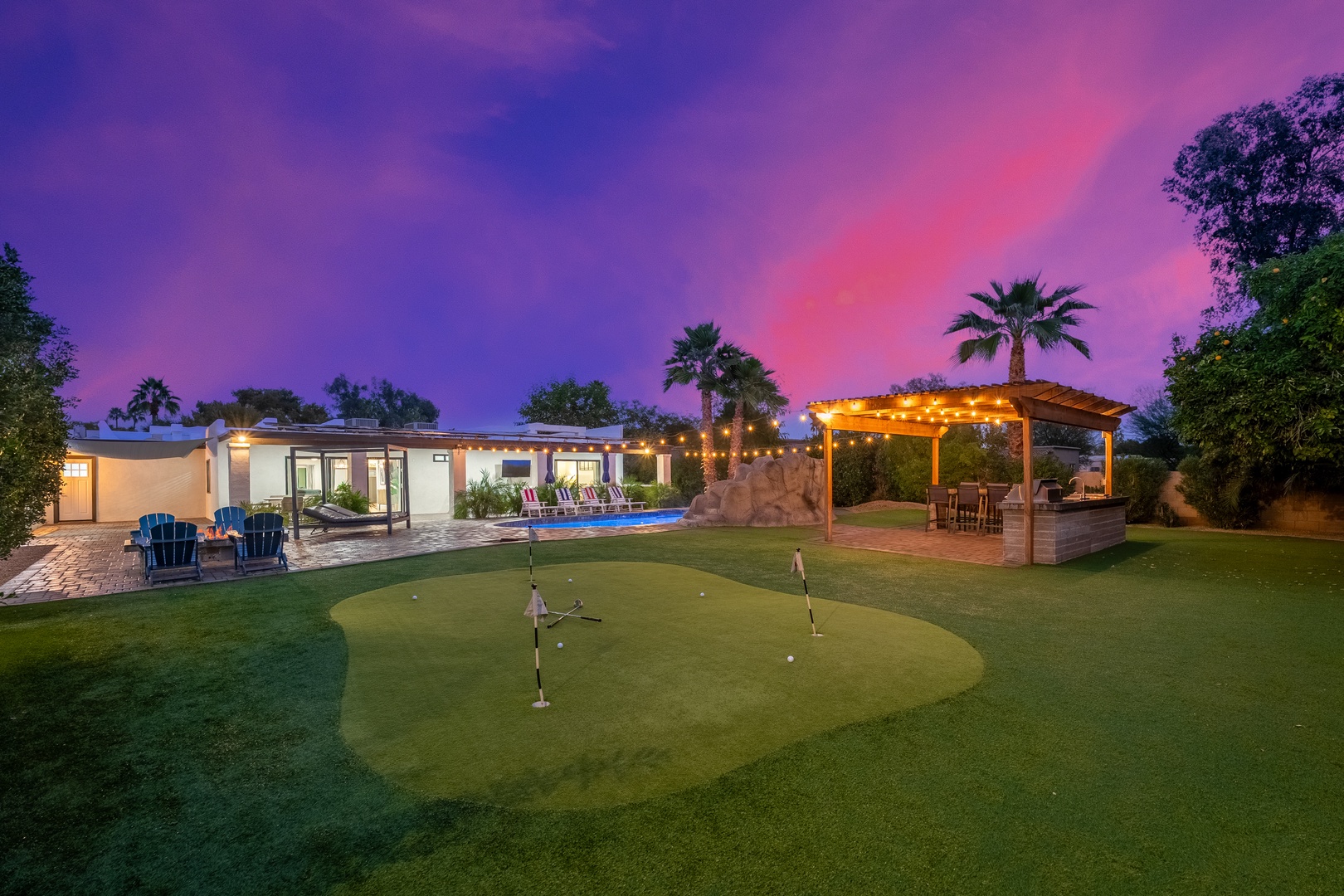 Stunning sunset paints the sky in vibrant purples and pinks above the property, featuring a private putting green, illuminated pool, and covered patio perfect for relaxing evenings.