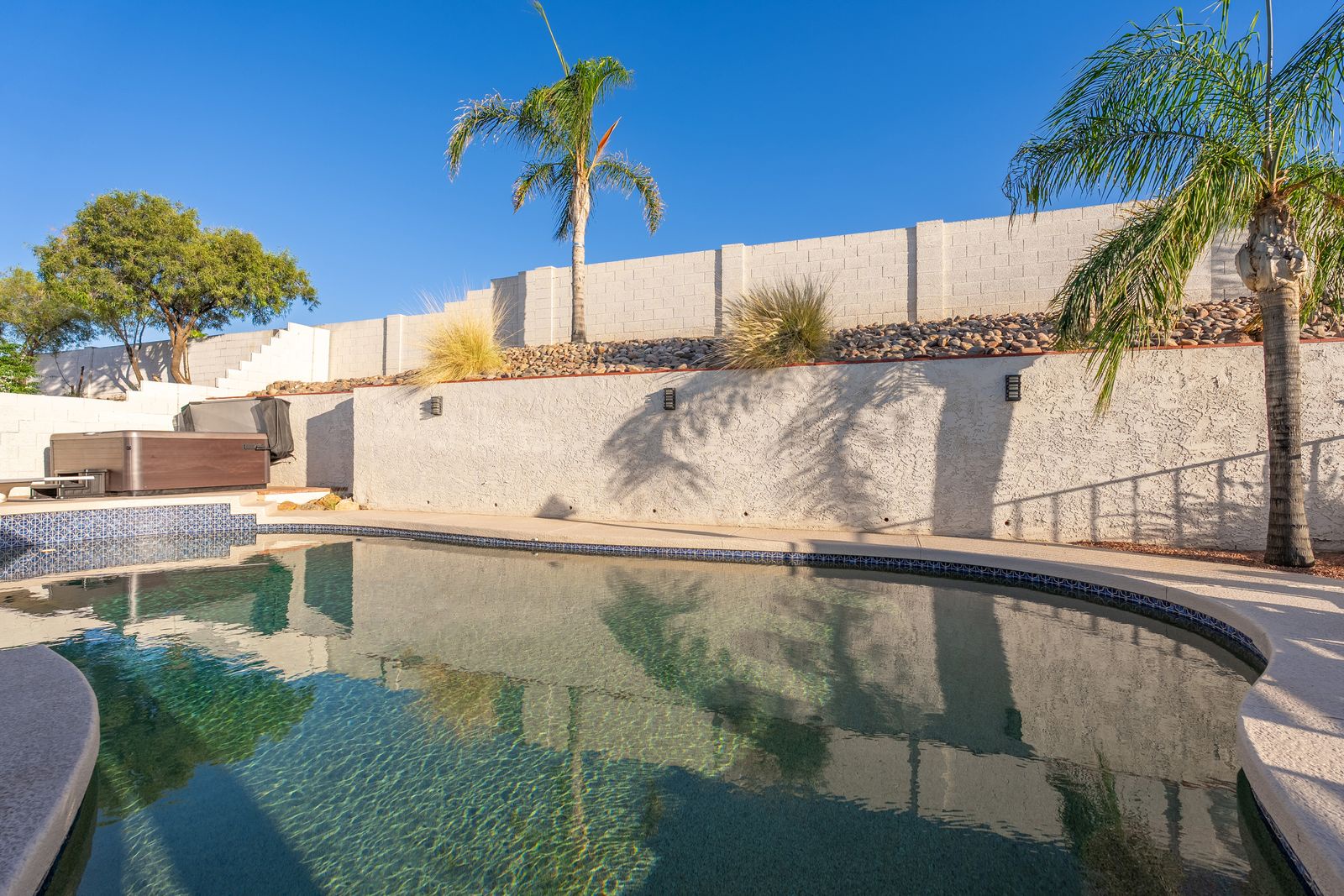 Private backyard oasis featuring a sparkling pool surrounded by swaying palms, warm desert landscaping, and a covered lounge area—your perfect escape under endless blue skies.