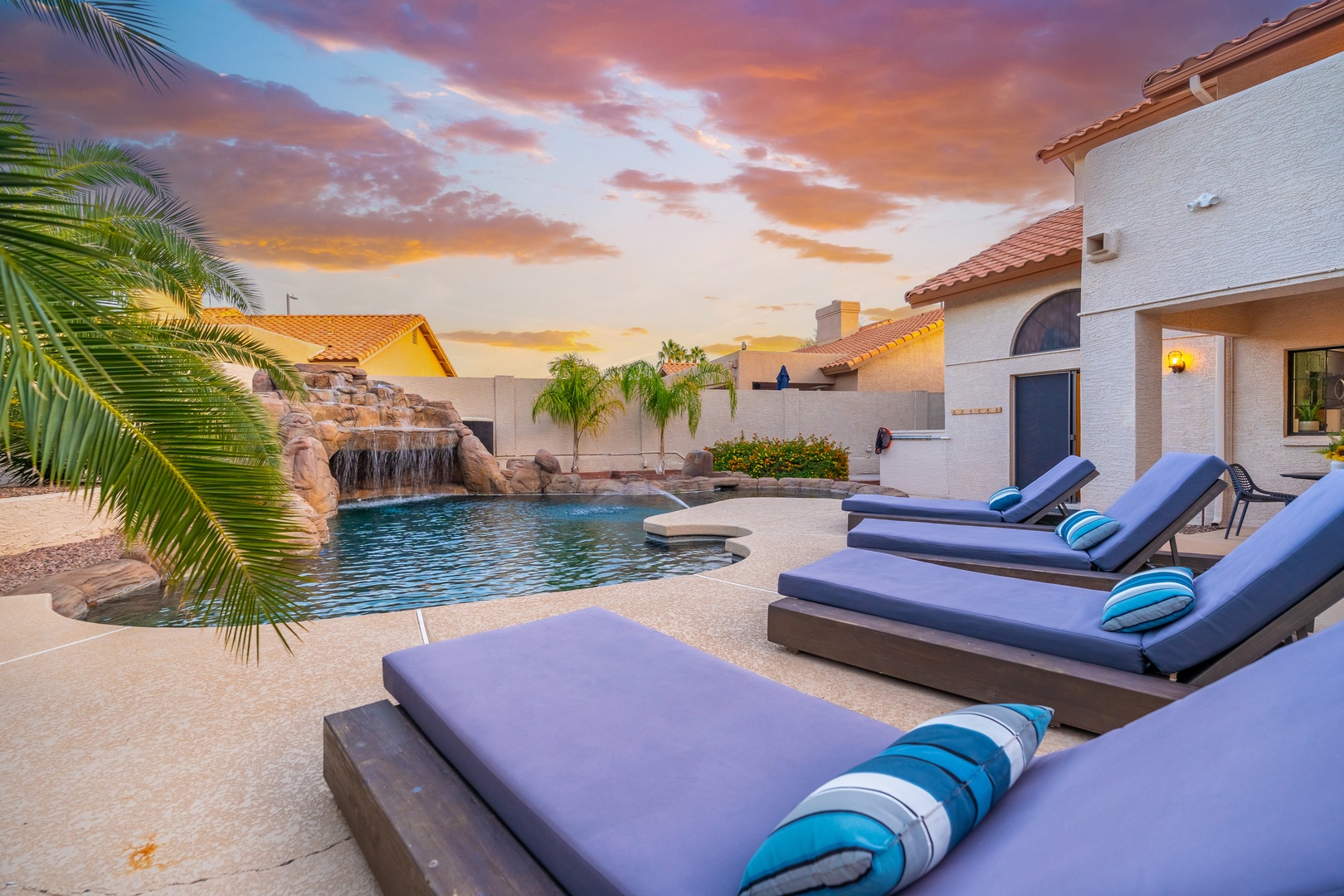 This backyard retreat features a sparkling pool with a cascading rock waterfall, surrounded by comfortable loungers. The property showcases southwestern architecture with terracotta roofing and desert landscaping beneath colorful skies.