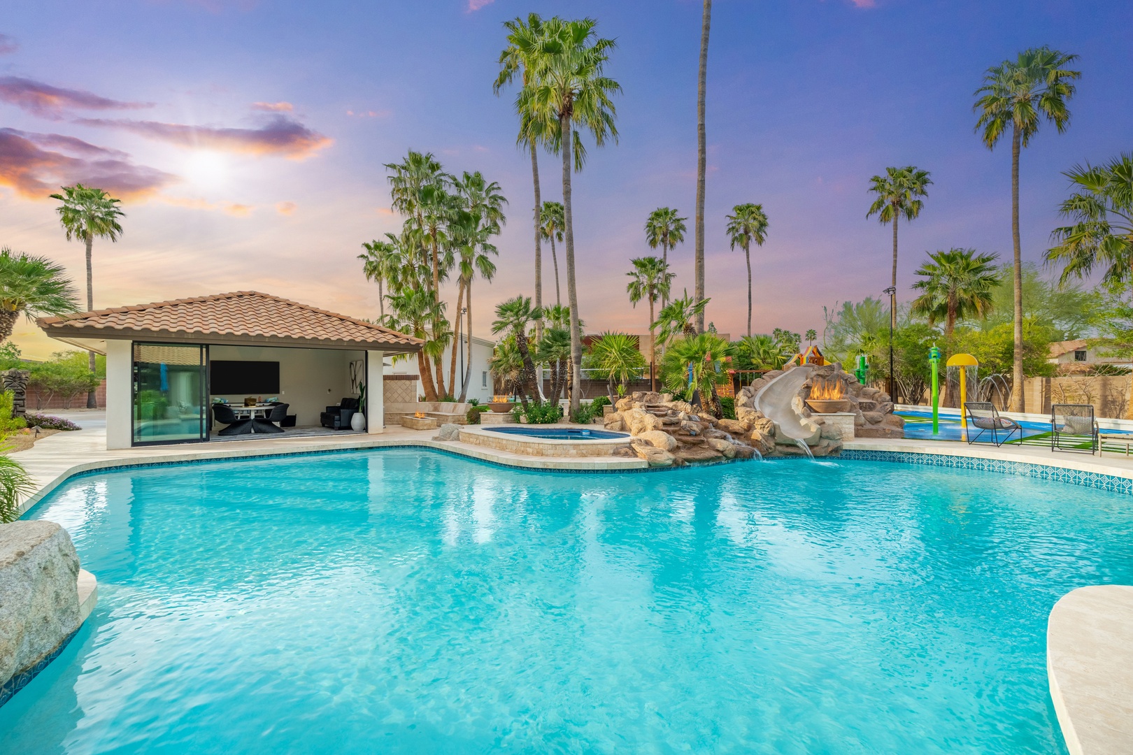 An expansive tropical oasis showcasing a sparkling pool with waterslide, swaying palms, and a shaded cabana area beneath a stunning painted sunset sky.