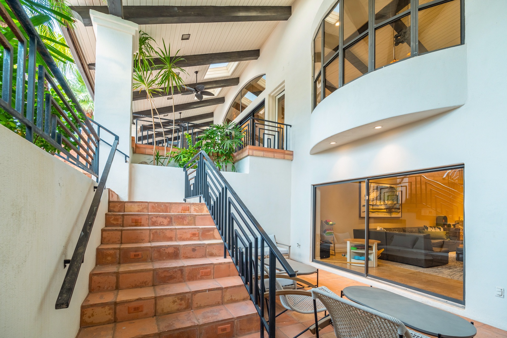 Sunlight streams through lush palms, illuminating your multi-level retreat's open staircase and vaulted ceilings. Exposed beams, curved balconies, and modern accents blend into a sophisticated, airy sanctuary inviting you upstairs.