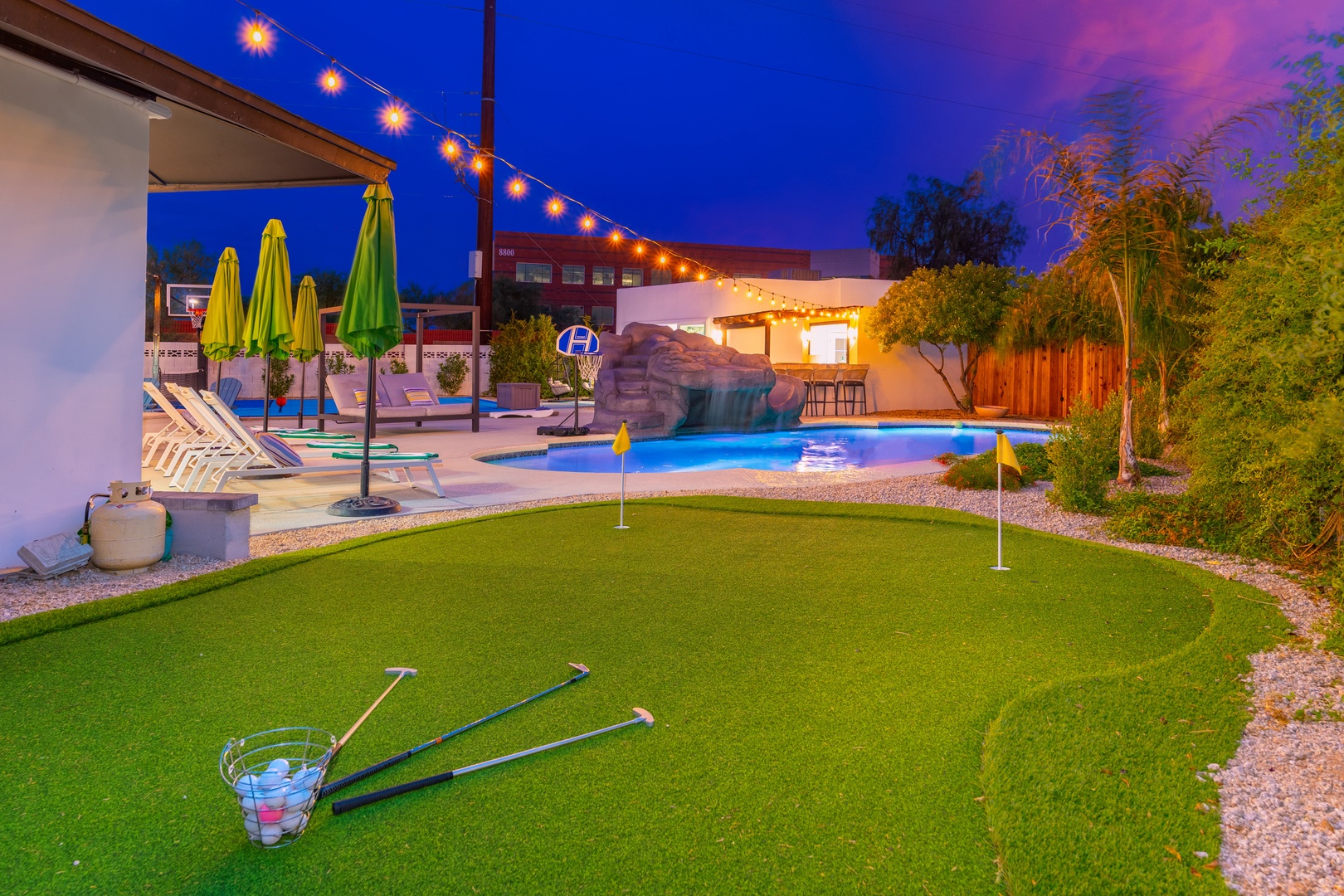 The property features a resort-style backyard with a mini golf green, cascading waterfall pool, spa, lounge seating under umbrellas, and festive string lights—all set against a stunning evening sky.