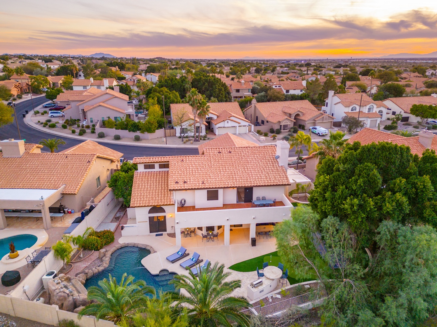 Aerial view captures a residential neighborhood at sunset, showcasing the property's distinctive architecture and surroundings with mountains visible in the distance.
