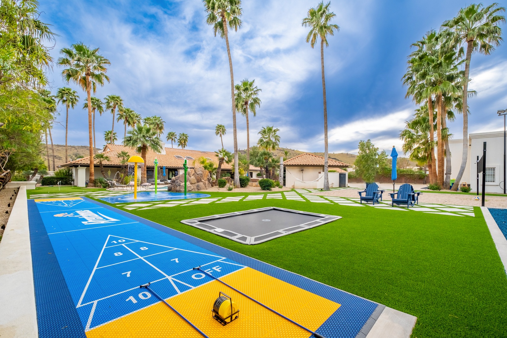 A spacious outdoor recreation area featuring hopscotch, trampolines, and vibrant blue seating beneath towering palm trees, framed by desert hills and sunny skies.