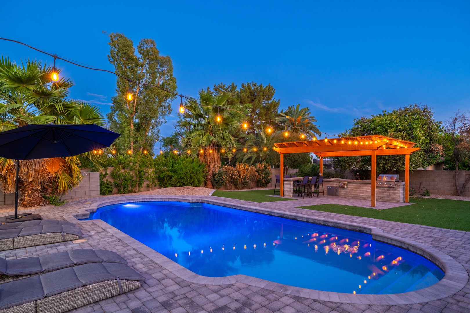 A private backyard oasis at dusk, featuring a sparkling pool, covered patio dining area, and tropical landscaping illuminated by string lights beneath a vibrant blue evening sky.