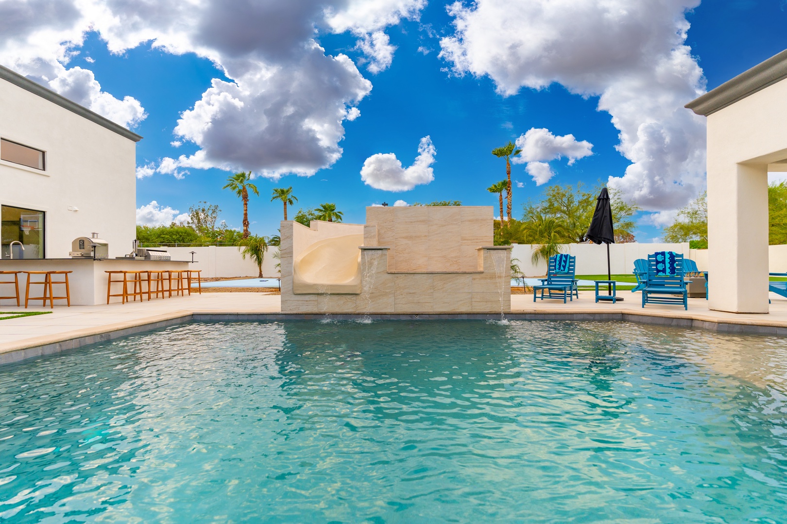 A stunning outdoor oasis awaits with a sparkling turquoise pool, modern waterfall feature, vibrant lounge seating, and desert palms beneath bright blue skies with fluffy white clouds.