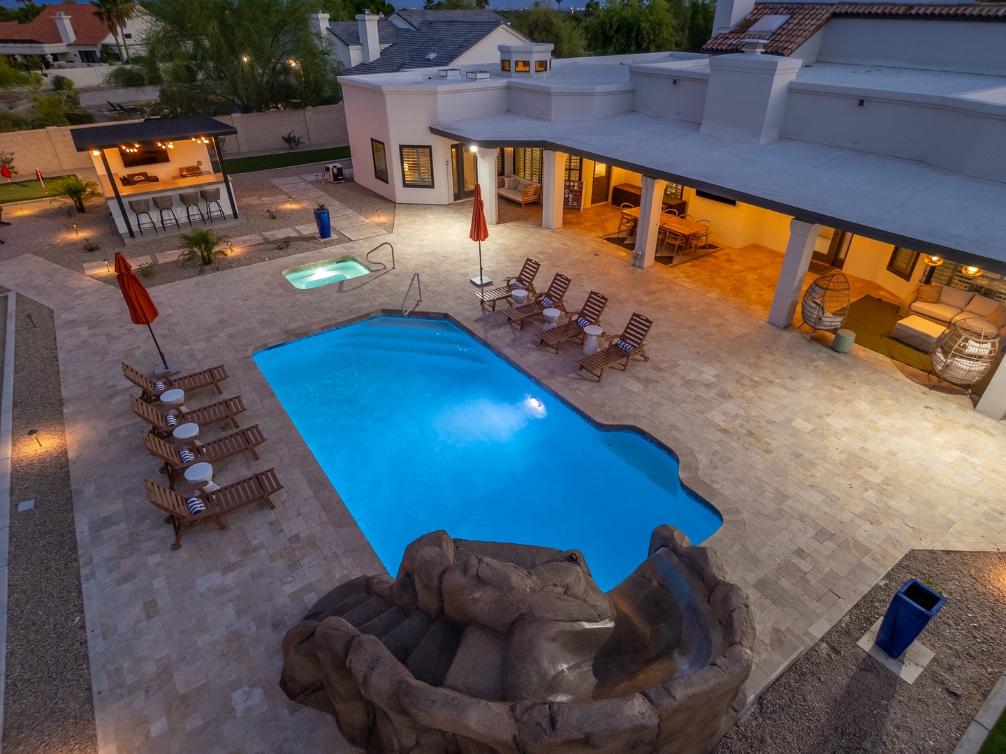 Aerial view of the property showcasing an expansive pool area, a separate hot tub with rock-inspired design, covered outdoor lounge spaces, and a well-appointed backyard in a quiet residential neighborhood.