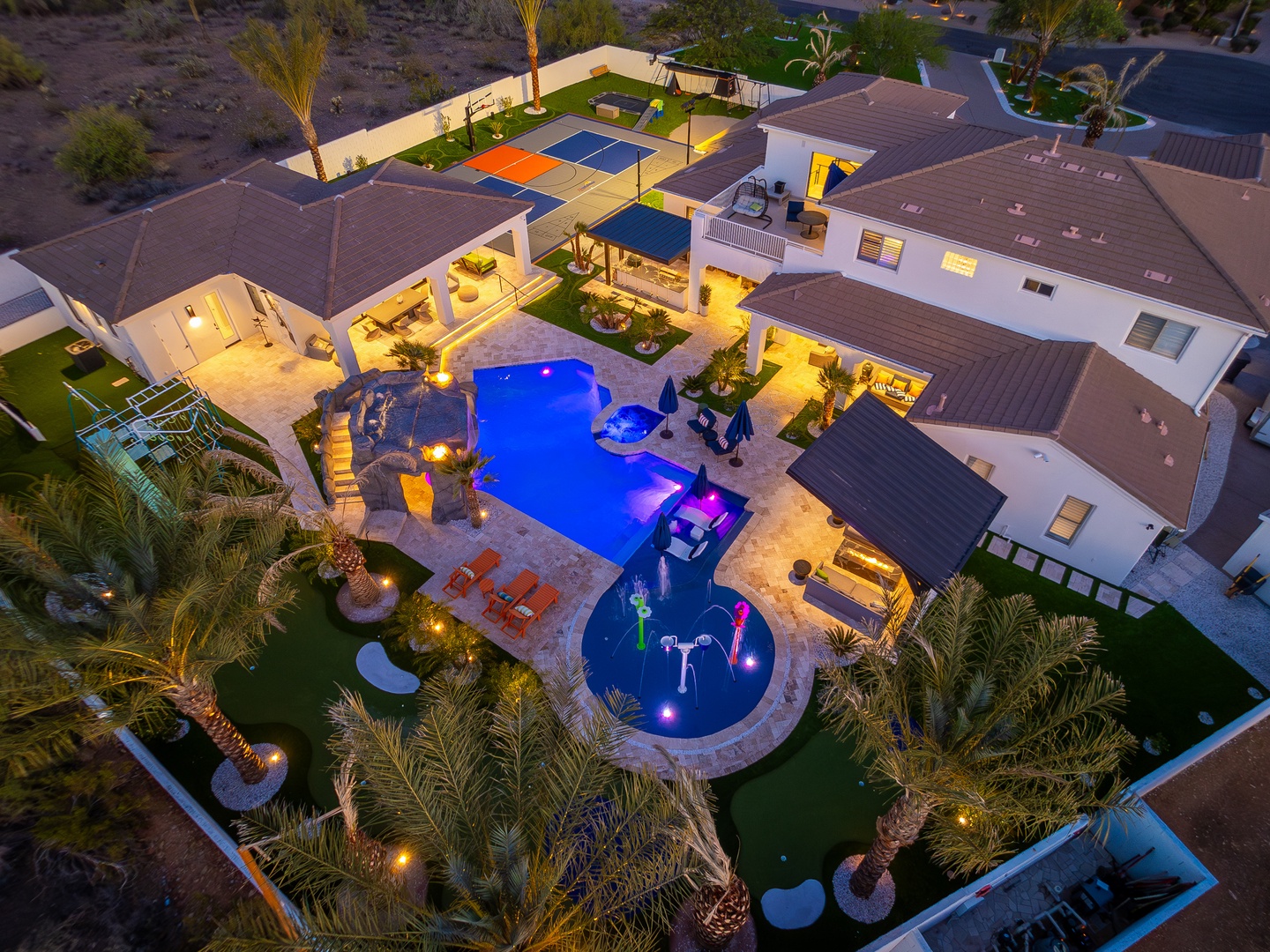 Aerial view of a luxurious desert vacation property featuring a stunning lagoon-style pool, sports court, and expansive outdoor living areas surrounded by palm trees and mountain landscape.