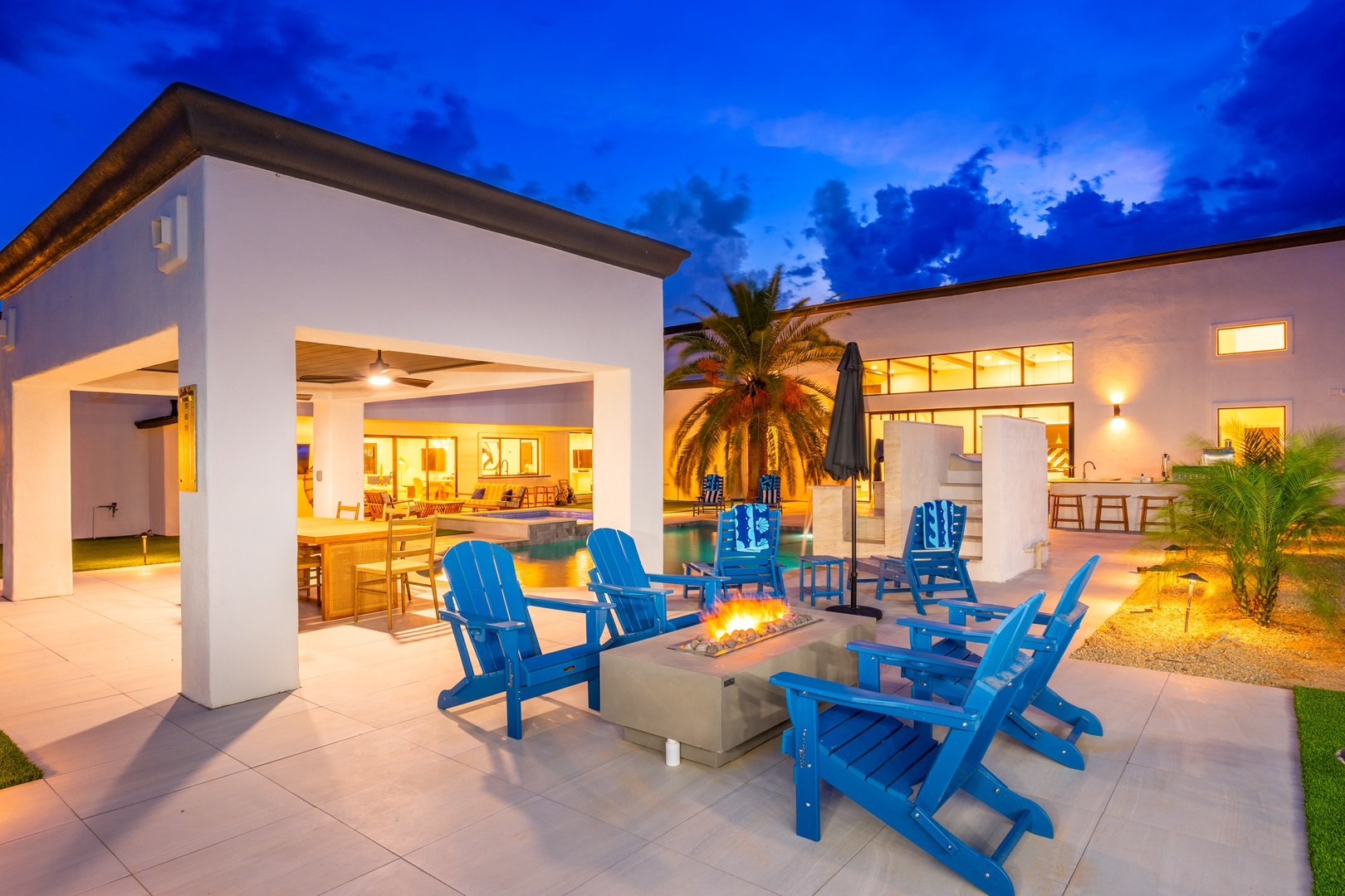 Modern desert oasis featuring a stunning outdoor living space at twilight. Vibrant blue seating encircles a contemporary fire feature beneath an open-air pavilion, while the pool's glow invites relaxation as dusk settles in.