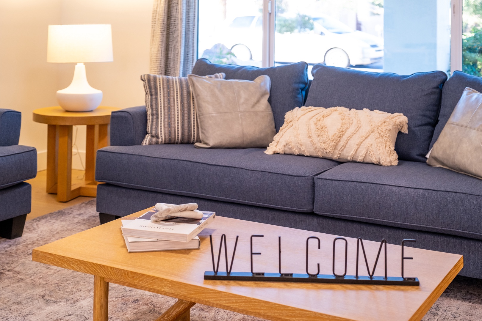 Sink into plush cushions on your stylish gray sofa, where natural light fills your living space through bright windows. Thoughtful touches like soft throw pillows and a welcoming sign set the tone for your stay.