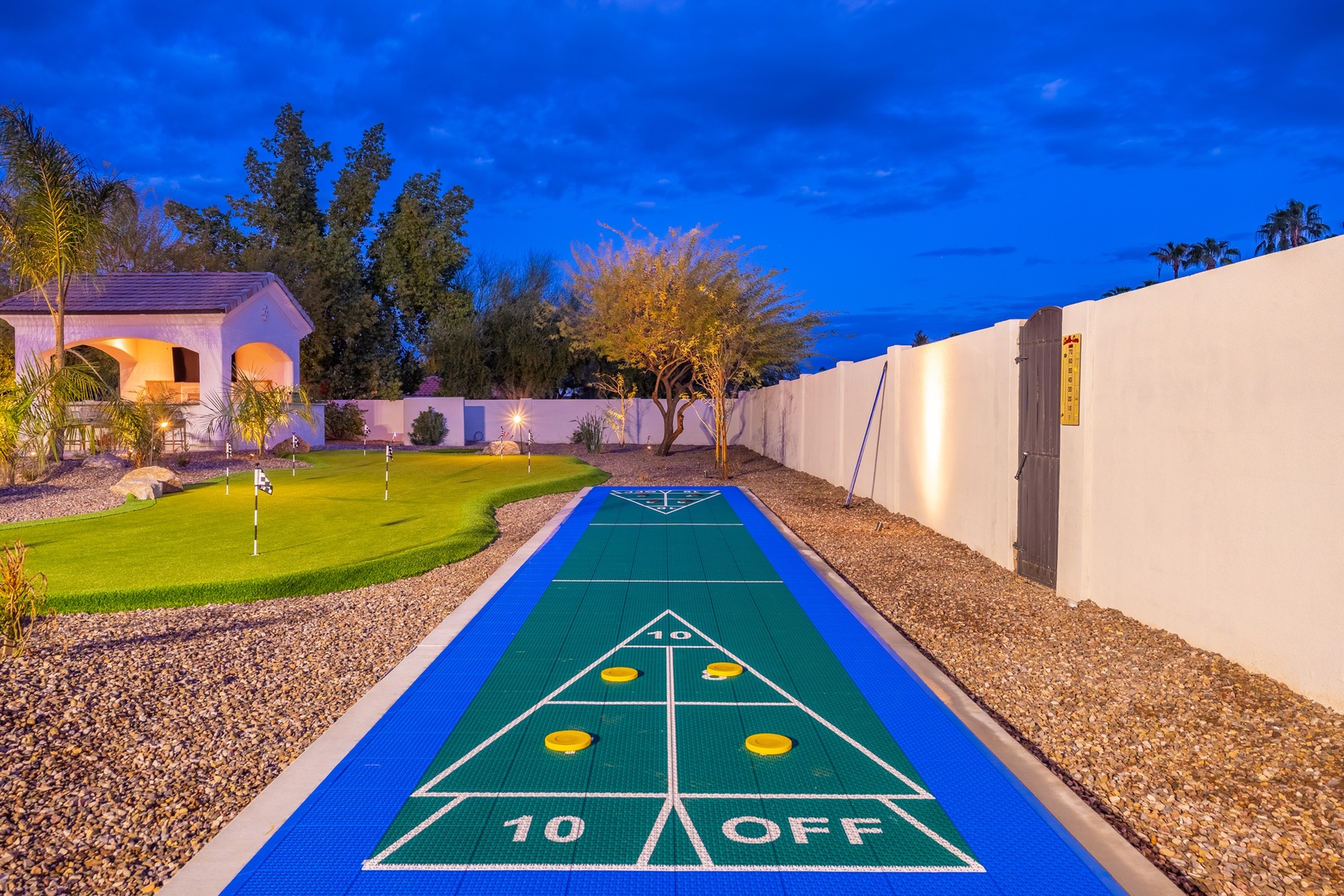 A private outdoor shuffleboard court and putting green glowing under evening lights, surrounded by desert landscaping and palm trees, perfect for friendly competition and relaxation.