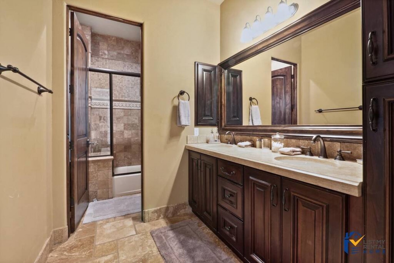 Prepare for your day in this elegant bathroom featuring rich wood cabinetry, dual vanities, and thoughtful amenities for a luxurious start.