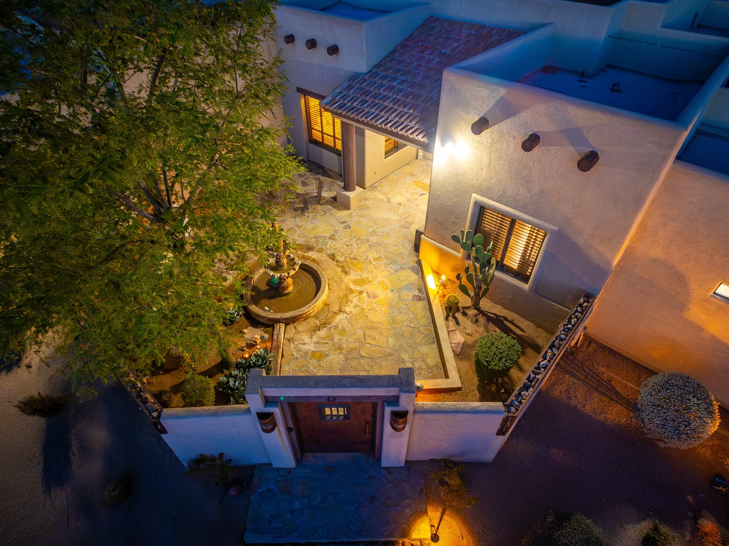 Aerial view of the property at twilight showcases charming Mediterranean architecture, a central courtyard fountain, lush landscaping, and warm ambient lighting throughout the outdoor spaces.