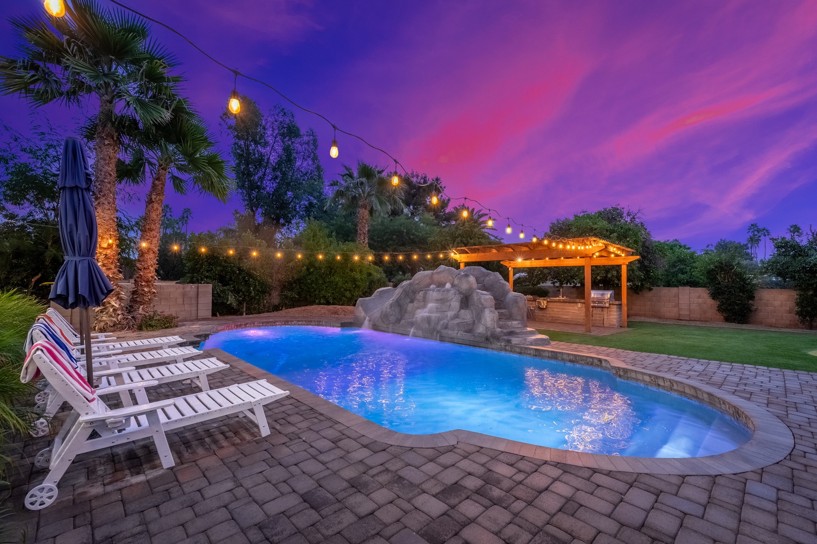 A backyard oasis featuring a sparkling pool with waterfall rocks, paver patio, and covered pergola beneath a stunning purple twilight sky.