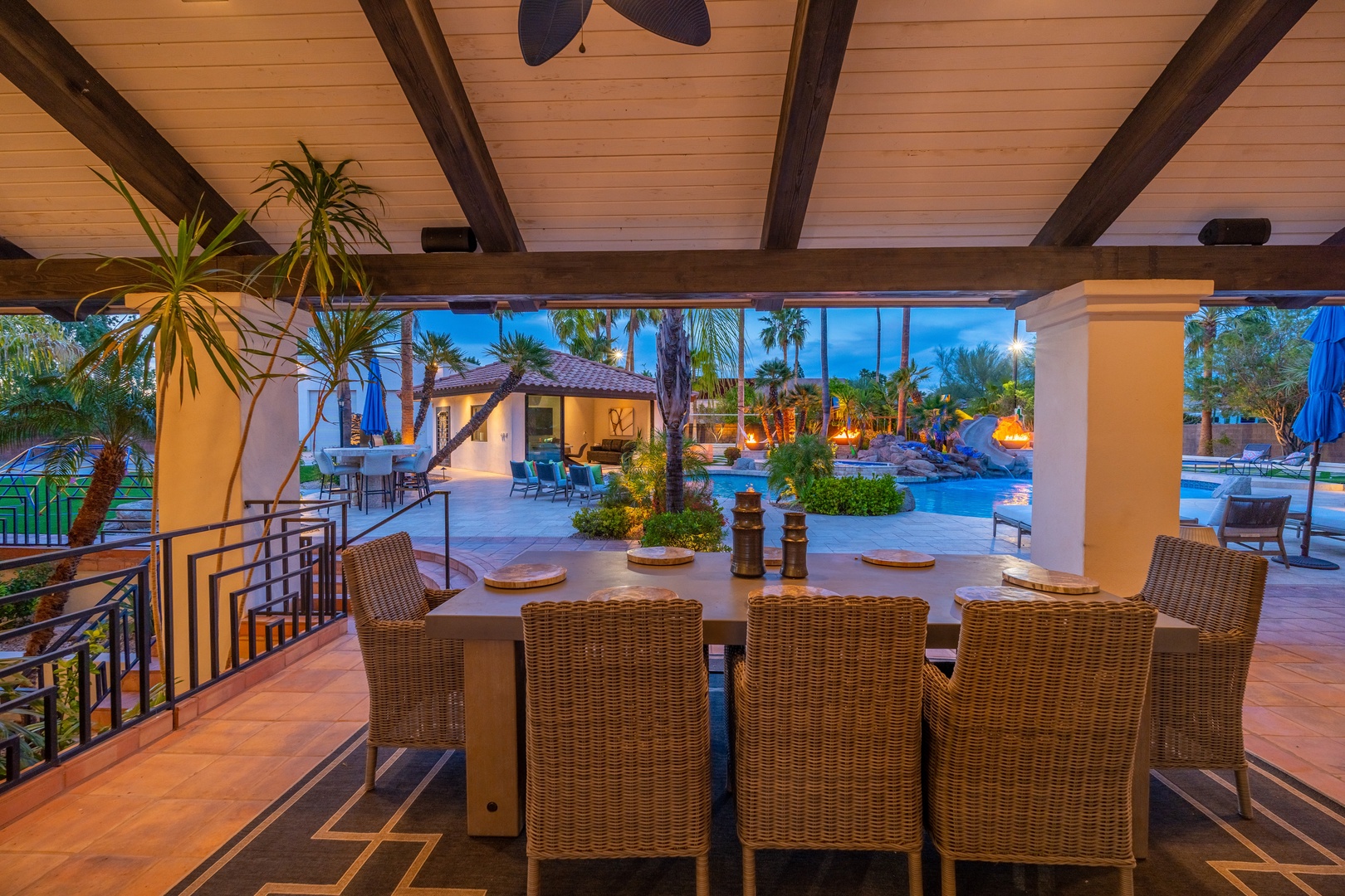 The exterior structure of the property at dusk, showcasing a covered outdoor dining area with pool and palm-lined grounds beyond, all framed by warm architectural lighting.