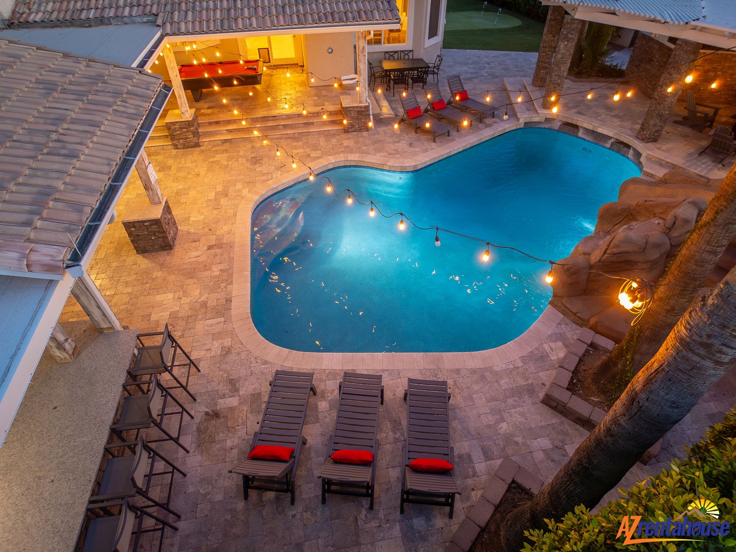 Aerial view of a beautifully lit villa featuring a resort-style pool, spacious patio with lounge chairs, and outdoor seating areas perfect for evening relaxation and entertaining.
