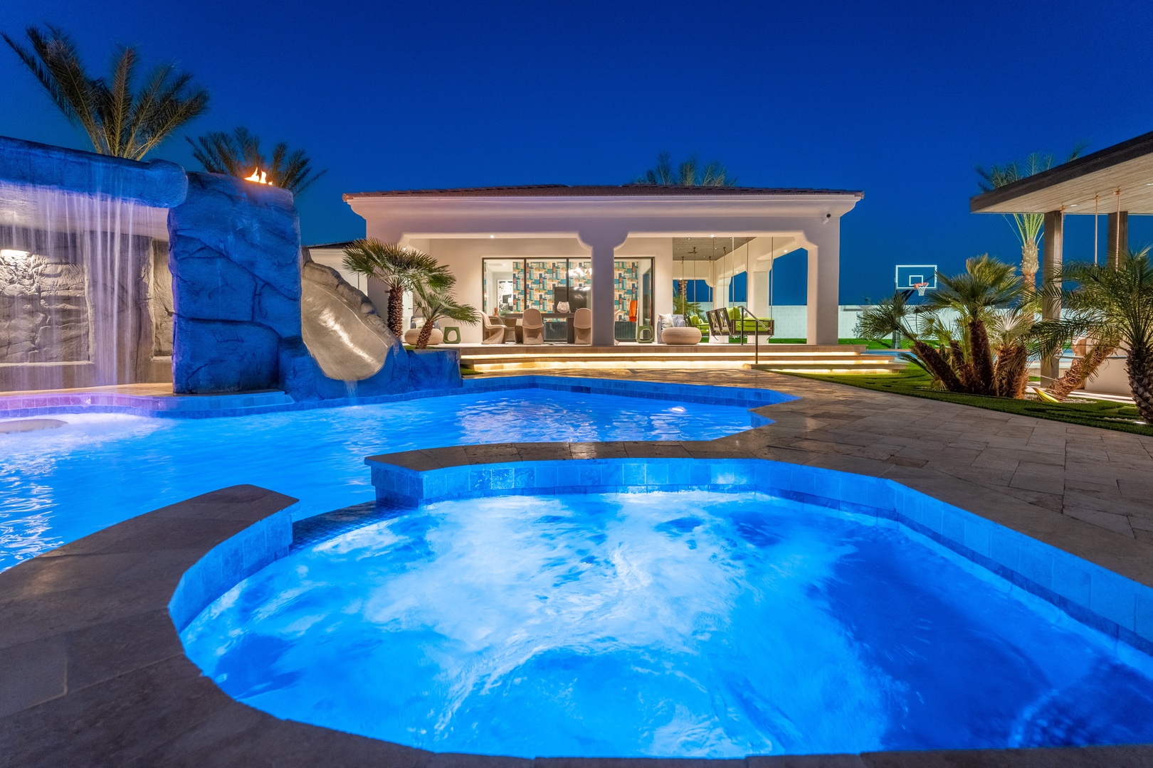 This modern villa's pool area glows with ambient lighting as the evening sets in, showcasing a luxurious hot tub, waterslide, and spacious open-air entertaining spaces perfect for memorable vacation moments.