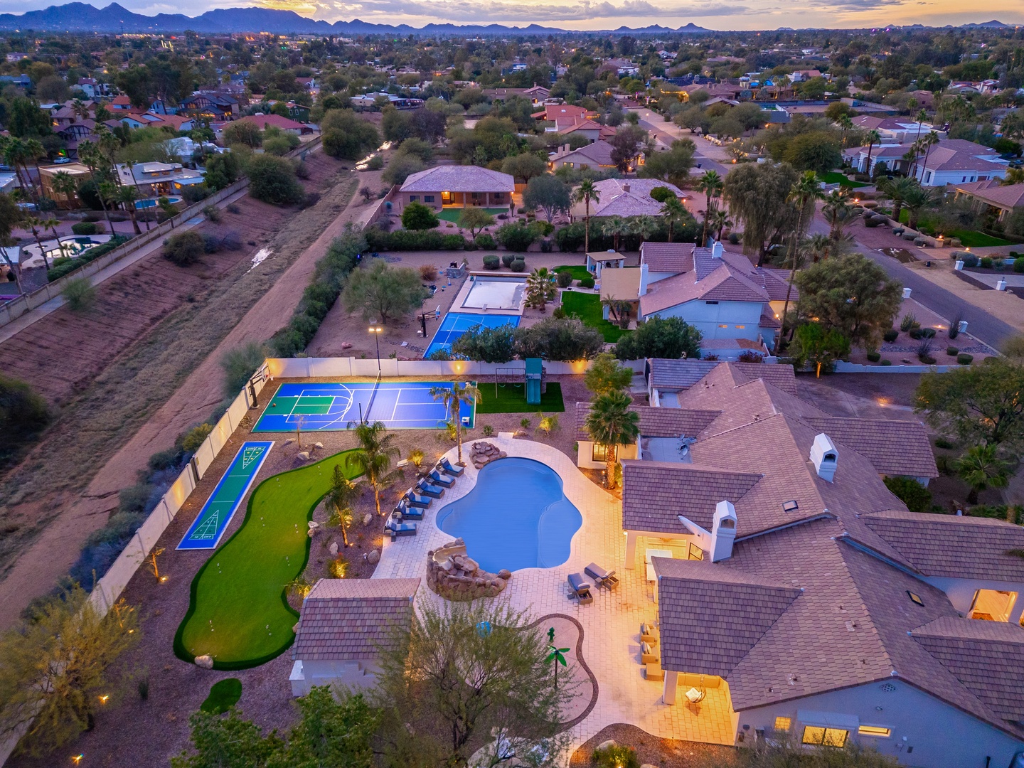 Aerial view of a sprawling resort-style property with swimming pools, tennis court, mini golf, and mountain backdrop in a tranquil desert neighborhood at twilight.
