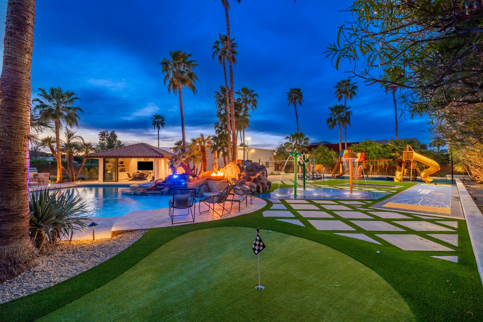 Expansive vacation estate at dusk, featuring a resort-style pool, playground, putting green, and tropical landscaping under towering palms and desert skies.