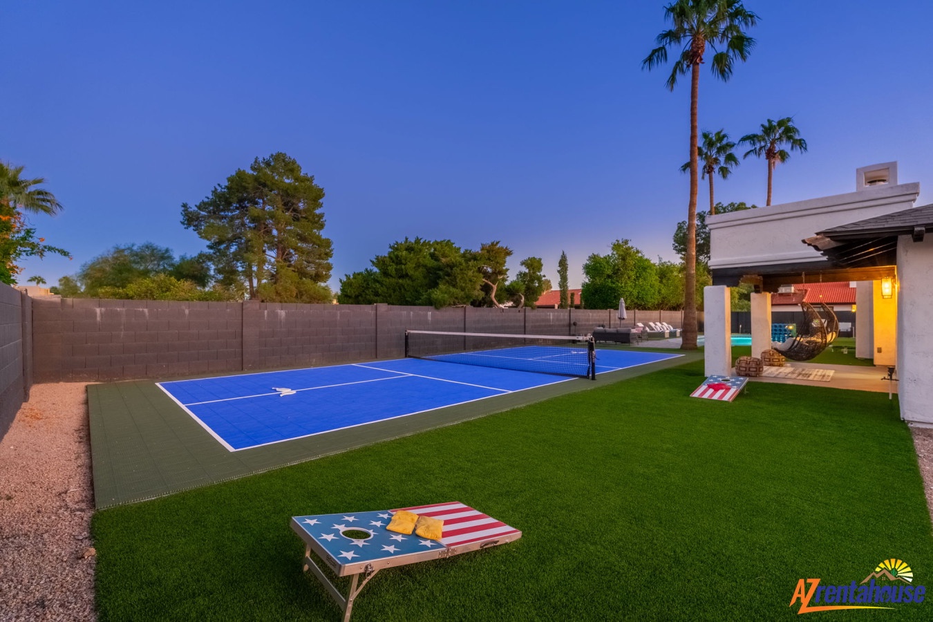 Private backyard features tennis court and game area with artificial turf, surrounded by palm trees and evening lighting.