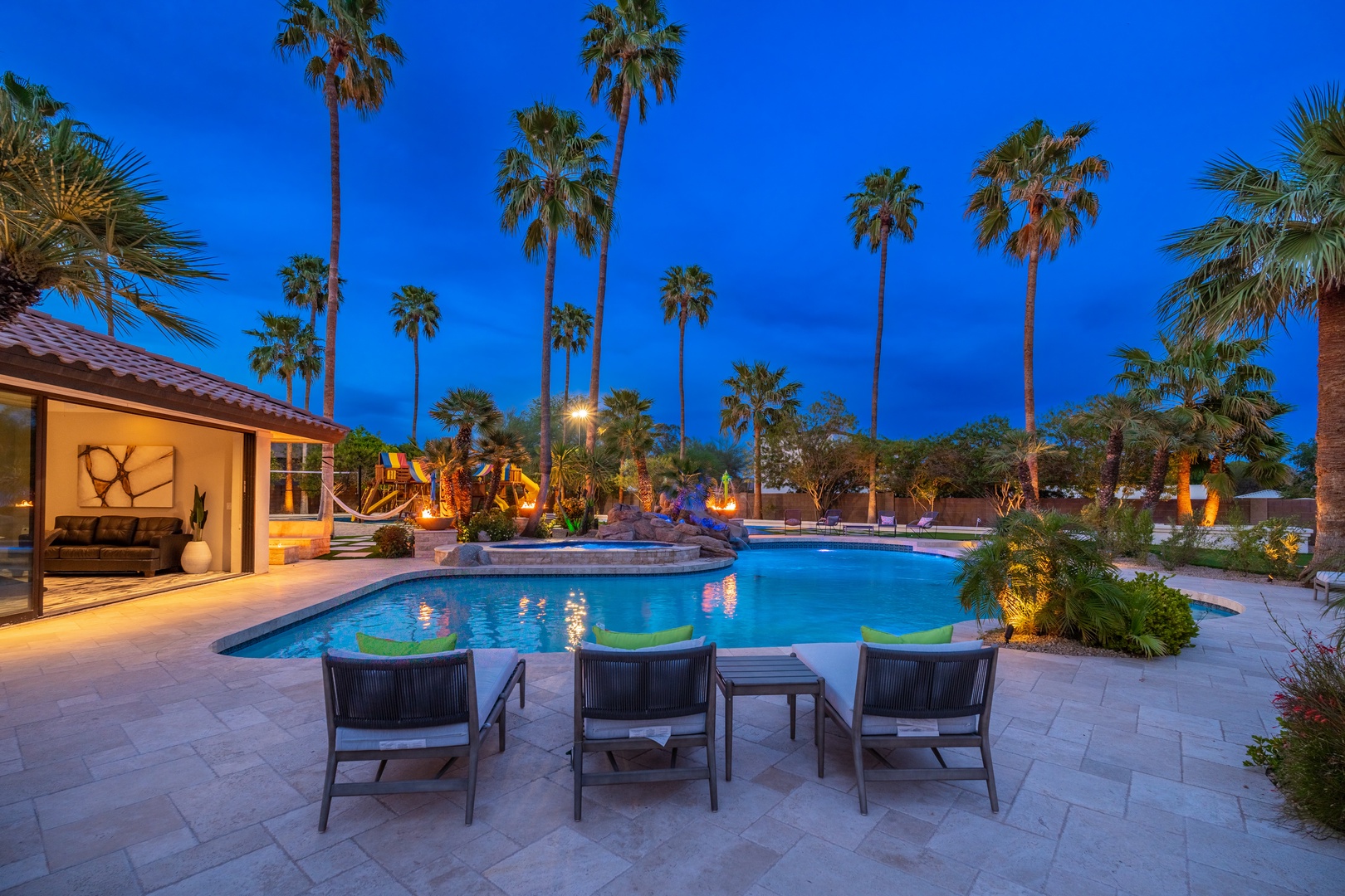 Stunning resort-style backyard retreat featuring illuminated pool, tropical palms, and lounge chairs perfect for evening relaxation under the desert sky.