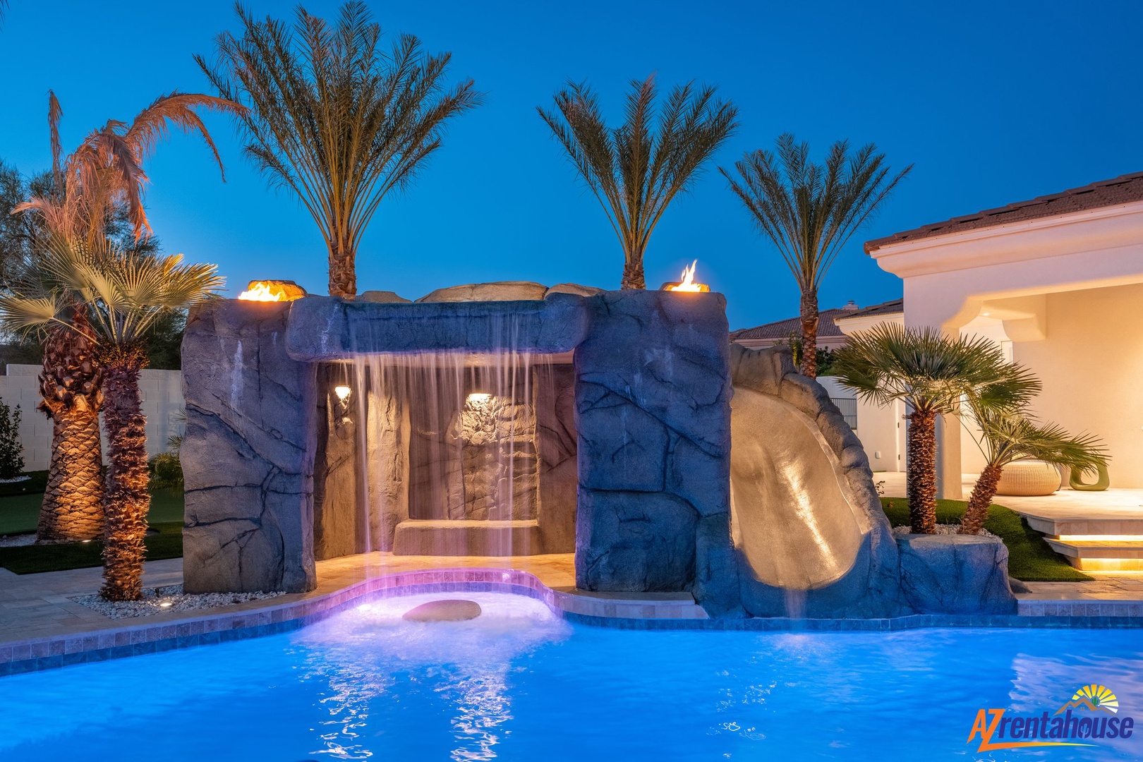 Stunning resort-style pool with waterfall, slide and spa under evening lights surrounded by palm trees.