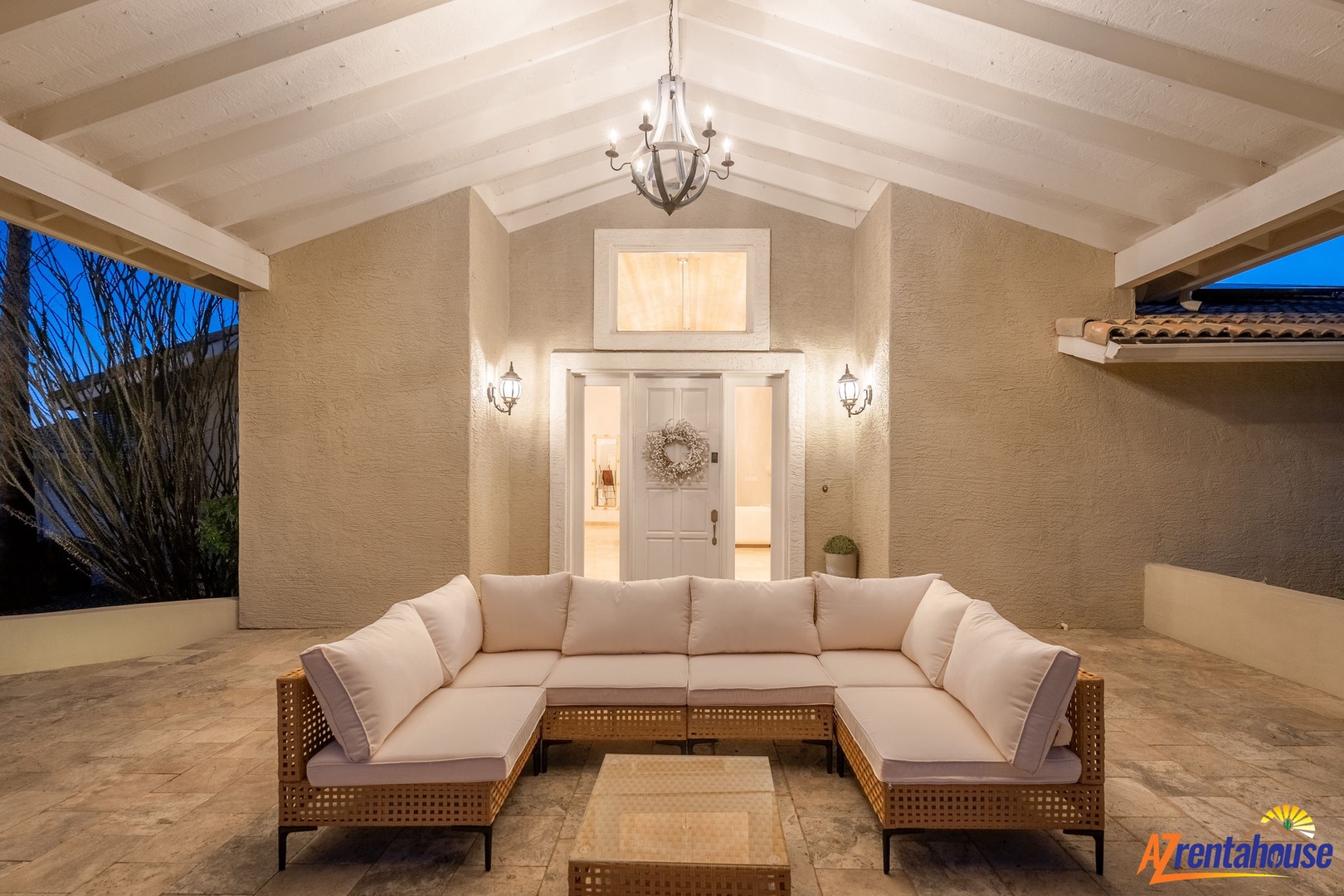 A beautifully lit covered patio awaits your evening conversations, with plush seating beneath soaring vaulted ceilings and elegant chandelier glow as twilight sets in.