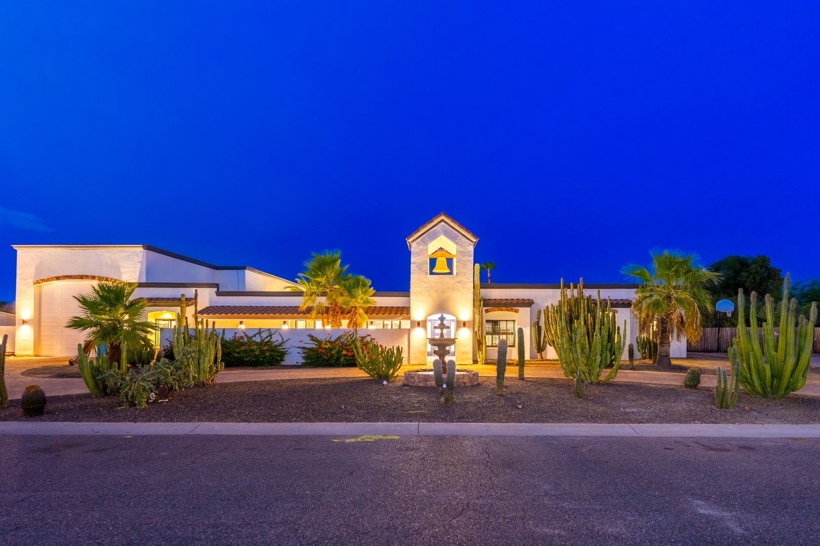 A striking Spanish-style property with a distinctive bell tower entrance, surrounded by mature cacti and palm trees in a desert landscape, photographed at dusk.