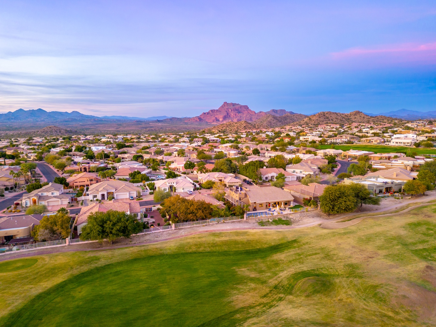 Aerial view of a residential neighborhood nestled against dramatic red rock mountains, captured during the soft twilight hour with pink and blue hues painting the desert sky.