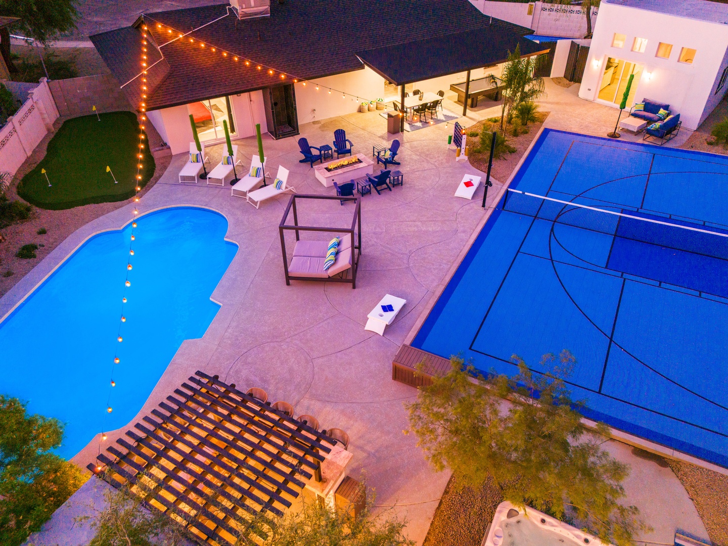 Aerial view of a modern vacation home at dusk featuring a sparkling pool, full-size tennis court, outdoor dining and lounge areas, and illuminated patio spaces.