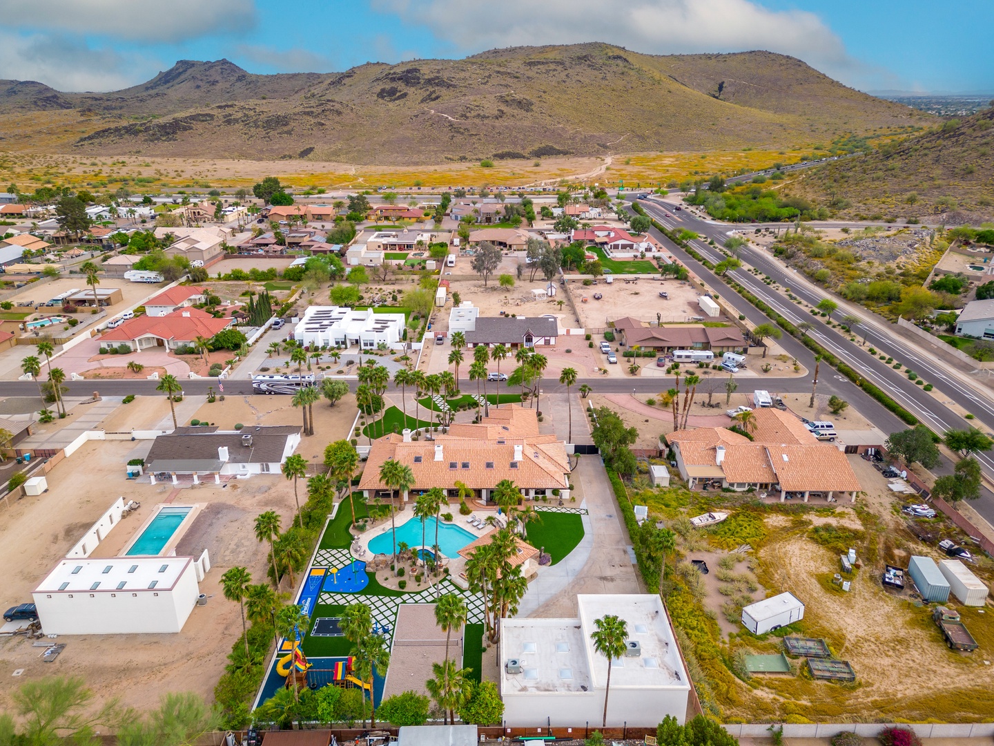 Aerial view of a desert neighborhood property featuring two pools surrounded by palm trees and recreational areas, set against dramatic mountain landscapes and wide-open skies.