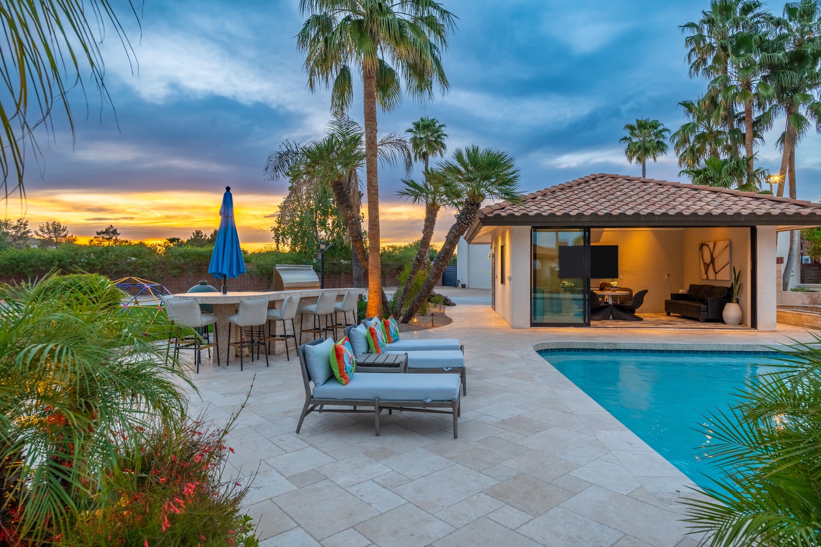A private desert oasis awaits beneath swaying palms, where stunning sunset views frame the shimmering pool and expansive patio—the perfect setting to unwind with colorful cushioned loungers and an inviting outdoor bar.