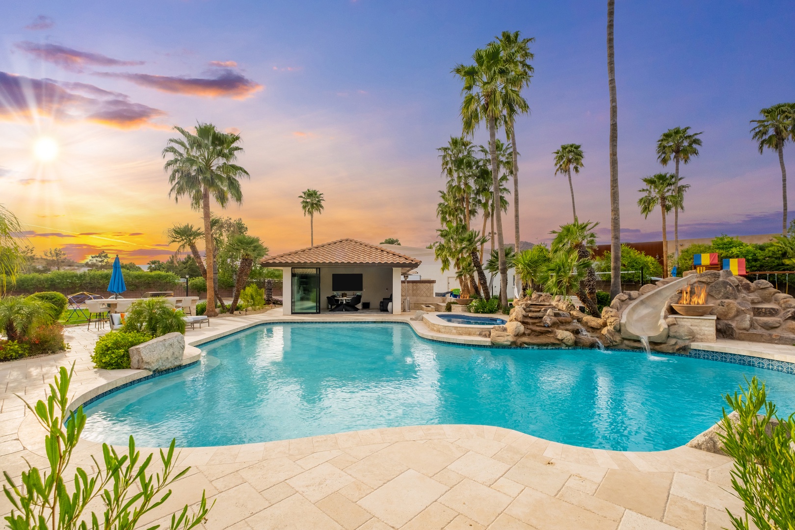 The property features a resort-style pool with waterfall and slide, surrounded by mature palm trees and lush landscaping, with covered patio area and stunning desert sunset views.