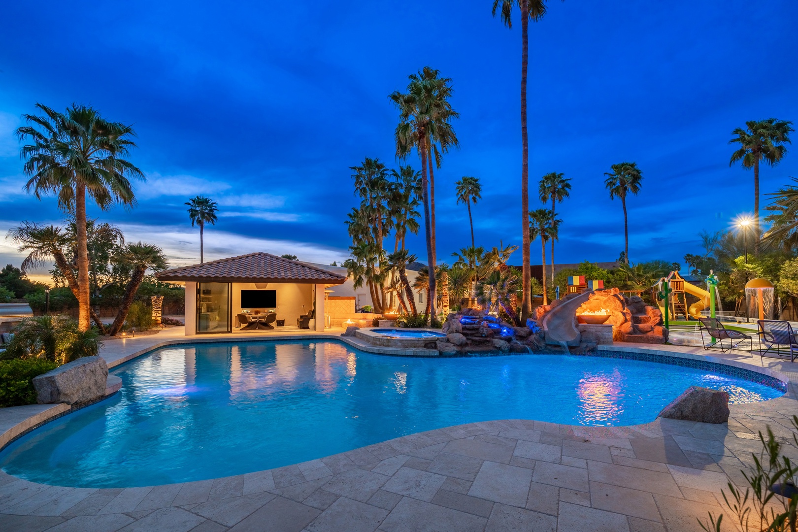 A stunning desert retreat featuring a custom freeform pool with natural rock formations, hot tub, waterslide, and towering palms framing a vibrant twilight sky.