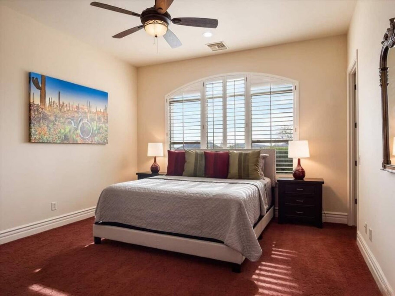 Relax in your peaceful bedroom with a plush bed, vibrant desert artwork, and cooling ceiling fan. Large plantation shutters frame natural light while colorful accents add warmth to your desert retreat.
