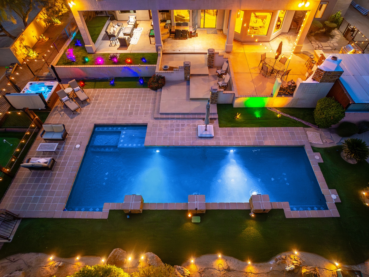 Aerial view of the illuminated outdoor recreation area featuring a large swimming pool, multiple lounging spaces, and architectural highlights of the vacation rental at night.